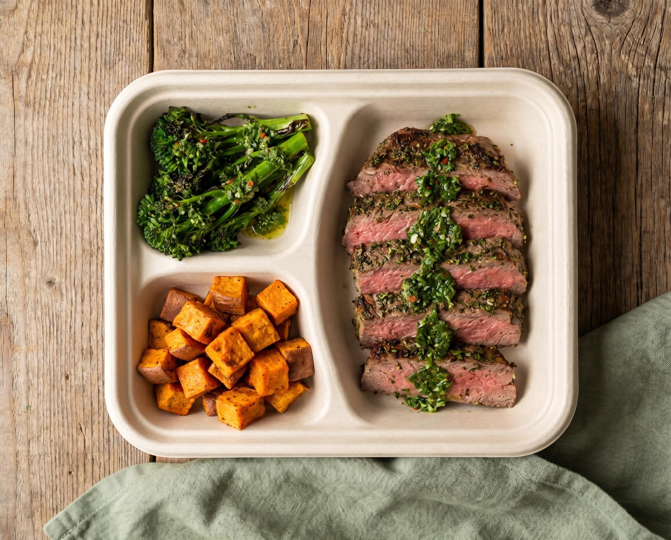 Three-compartment meal prep tray with grass-fed steak, chimichurri, roasted sweet potato, and broccolini